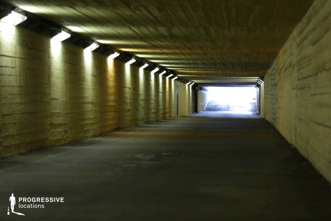 This image showcases a long, low-ceilinged concrete underpass with a repetitive pattern of overhead lights casting a moody, yellow-green glow along the textured walls. The tunnel extends into the distance, leading to a bright exit that contrasts sharply with the darker interior. The rough, unfinished concrete surfaces and artificial lighting create a stark, industrial atmosphere. This setting is ideal for filming tense urban dramas, crime thrillers, or chase sequences, offering a confined, gritty aesthetic that enhances suspense and mystery.