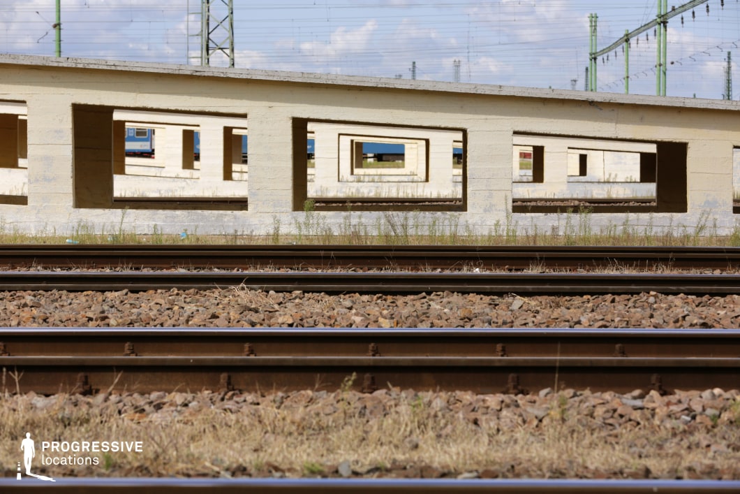 This industrial railway setting features multiple parallel train tracks running alongside a series of low, concrete structures with rectangular openings, creating a striking visual effect of depth and repetition. The rough-textured surfaces and geometric cutouts frame the background, where glimpses of rail infrastructure and distant trains are visible. Overgrown vegetation between the tracks adds to the sense of abandonment or decay. The strong linear composition and perspective make this location well-suited for filming tense chase sequences, crime dramas, or dystopian and post-apocalyptic narratives that emphasize desolation, industrial aesthetics, or transit-related themes.