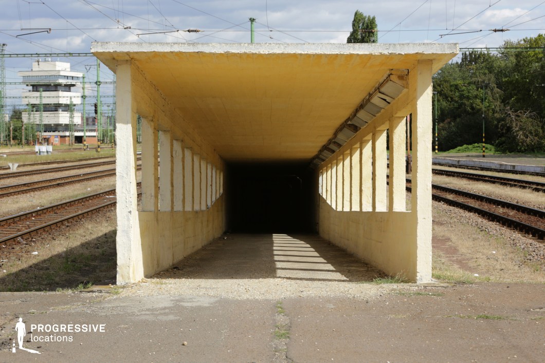 This railway underpass features a utilitarian, rectangular concrete structure with a weathered yellow finish. The repetitive vertical openings along the sides allow natural light to cast strong geometric shadows on the ground, creating a striking visual effect. The tunnel’s dark interior contrasts sharply with the bright exterior, enhancing the sense of depth and mystery. The surrounding railway tracks and distant control tower reinforce the industrial, transit-oriented atmosphere. This location is ideal for suspenseful or dramatic scenes in thriller, crime, or neo-noir films, serving as a setting for clandestine meetings, escape sequences, or moments of tension and uncertainty.