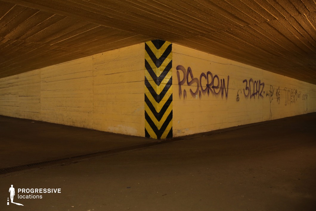 This image showcases a low-ceiling concrete underpass with yellow-painted walls adorned with graffiti, giving it an urban and gritty aesthetic. The corner is marked by a black-and-yellow striped hazard sign, reinforcing the industrial and utilitarian feel of the space. The rough texture of the concrete walls, coupled with the dim artificial lighting, creates a shadowy atmosphere, enhancing the sense of isolation and unease. The perspective leads into a vanishing point, making it visually compelling for dynamic shots. This location is well-suited for crime dramas, neo-noir thrillers, or underground subculture narratives, offering a raw, urban setting for clandestine meetings, chases, or introspective moments.