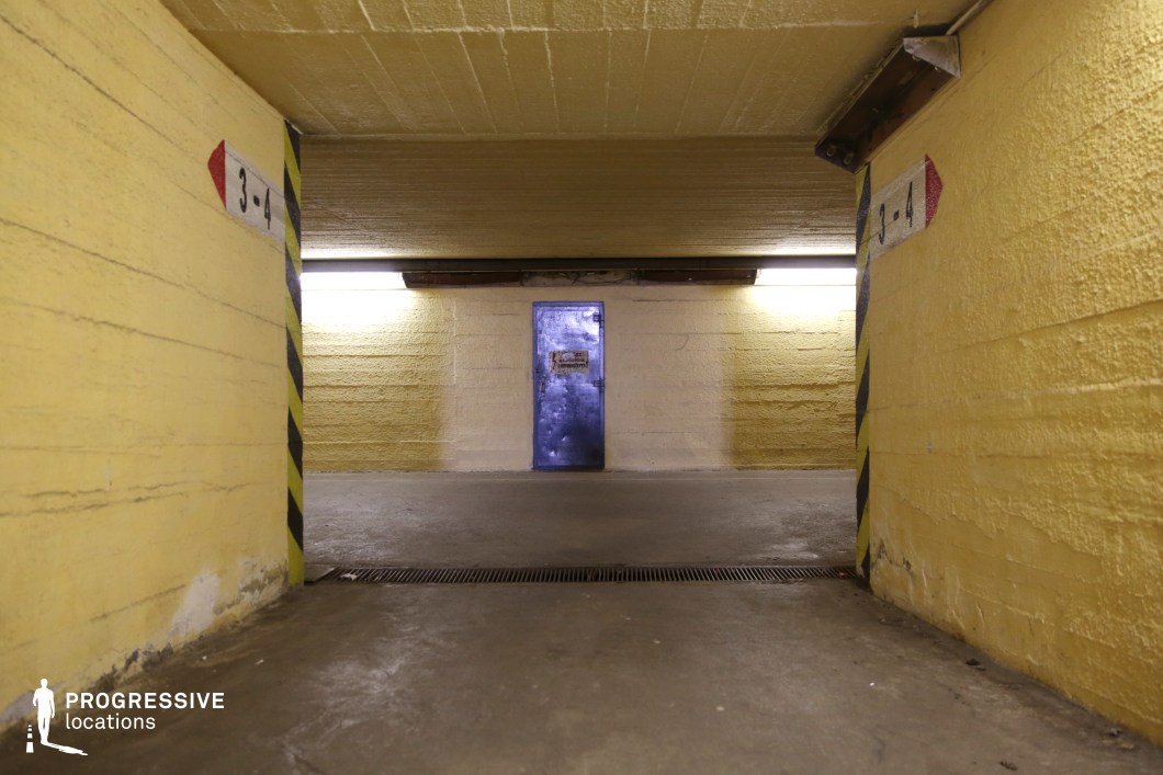 This confined underground passageway, lined with rough-textured yellow walls, leads to a striking metallic blue door at the far end, evoking a sense of secrecy and restricted access. The industrial aesthetic is reinforced by exposed structural elements, low ceilings, and functional strip lighting that casts a sterile glow. The striped hazard markings add an institutional or military feel, suggesting a former shelter, bunker, or subterranean maintenance facility. This location is ideal for filming tense or suspenseful scenes in dystopian, espionage, or sci-fi thrillers, serving as a clandestine meeting point, hidden research lab entrance, or high-security facility.