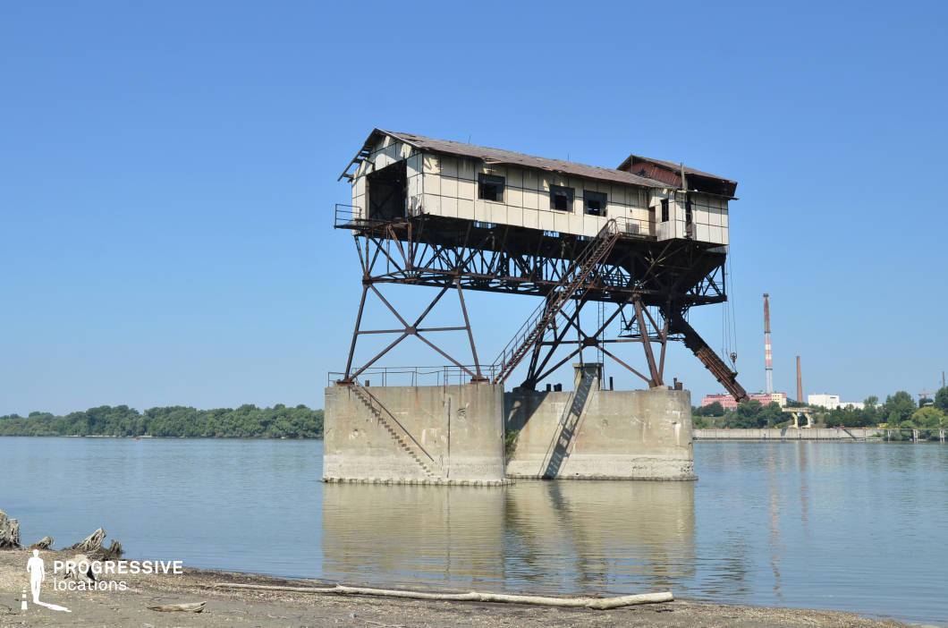 This abandoned industrial structure, precariously perched on rusted metal stilts above the water, exudes a haunting, post-apocalyptic presence. The weathered facade, broken windows, and steep metal staircase leading to the elevated platform create an atmosphere of decay and isolation. The concrete foundation, partially submerged, adds to the stark contrast between the structure and the surrounding natural landscape. In the background, distant smokestacks and industrial remnants reinforce the site's history of heavy industry. This location is ideal for dystopian films, survival thrillers, or crime dramas, offering a unique, desolate setting that conveys themes of abandonment and resilience.