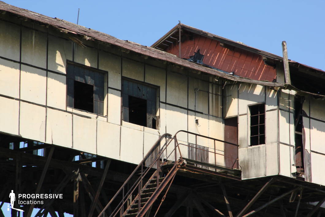 This decayed industrial structure, perched on a rusted steel framework, exudes an atmosphere of neglect and abandonment. The weathered exterior, with peeling paint, broken windows, and partially collapsed roofing, adds to the eerie and dystopian aesthetic. The metal staircase leading up to the entrance, combined with the exposed framework, enhances the precarious and desolate feel of the setting. This location is ideal for filming post-apocalyptic scenarios, crime thrillers, or urban exploration sequences, offering a gritty and atmospheric backdrop for tension-filled storytelling.