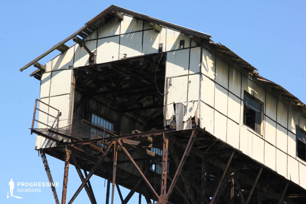 This deteriorating industrial structure, precariously perched on rusted metal supports, showcases extensive weathering with peeling panels, broken windows, and exposed wooden beams. The skeletal remains of the building reveal a dark, hollow interior, amplifying its abandoned and forgotten state. The corroded framework and makeshift repairs contribute to the scene’s rugged and unstable aesthetic. This location is ideal for post-apocalyptic films, industrial decay documentaries, or suspenseful thrillers, offering a raw, desolate, and ominous backdrop that evokes themes of abandonment and resilience.