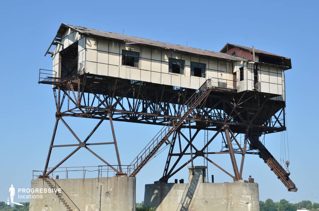 This eerie, elevated industrial structure stands precariously on a rusted metal framework, with a deteriorating wooden and panel exterior that shows signs of decay. The long staircase leading to the main platform adds a sense of height and isolation, while the exposed beams and skeletal supports emphasize its fragile, abandoned state. The industrial chute extending from the building suggests past functionality, likely as part of a loading or processing system. This location is ideal for dystopian films, post-apocalyptic settings, or crime thrillers, offering a gritty, atmospheric backdrop that evokes themes of decay, abandonment, and survival.