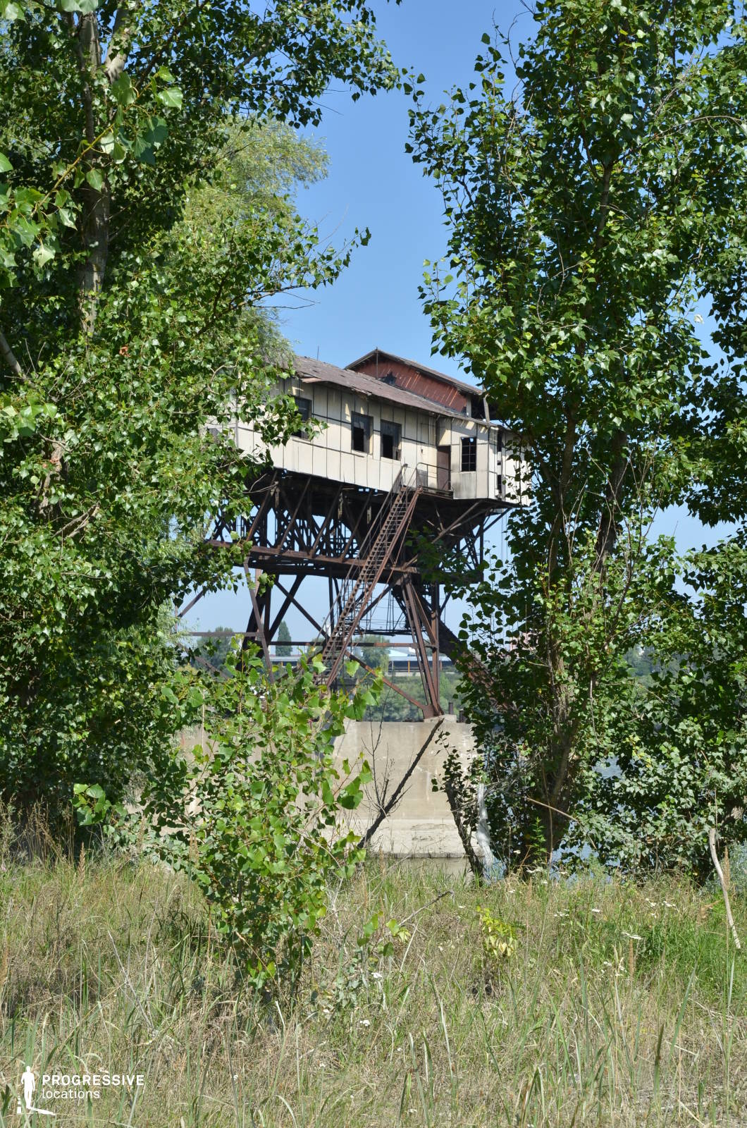 This elevated industrial structure, perched on rusted metal supports, stands in stark contrast to the surrounding lush vegetation. Its weathered facade, broken windows, and decayed metal framework suggest long-term abandonment, while the steep external staircase adds an element of precariousness. The dense greenery encroaching upon the site emphasizes a sense of nature reclaiming human-made structures. The location's mix of industrial decay and secluded wilderness makes it ideal for post-apocalyptic settings, survival thrillers, or mystery films, serving as a backdrop for hidden outposts, secret rendezvous, or eerie explorations of forgotten places.