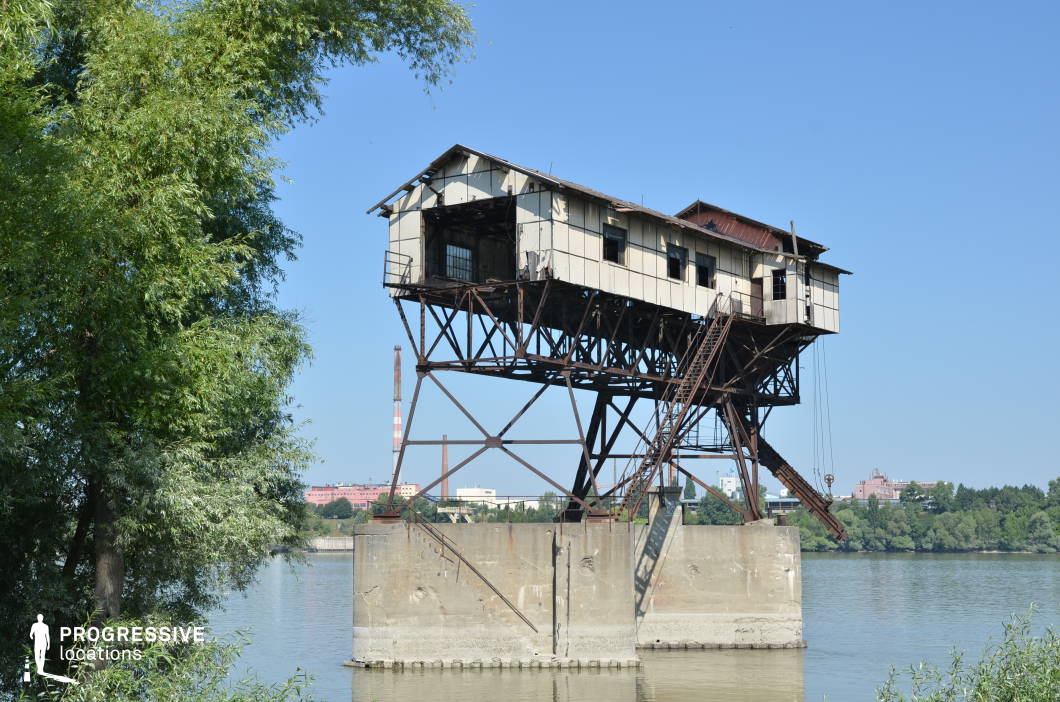 This decayed industrial building, precariously perched on rusting steel supports above the water, exudes an eerie, post-apocalyptic charm. The weathered metal framework and peeling paneling suggest years of neglect, while the steep external staircase adds an element of danger. Set against a backdrop of industrial chimneys and distant factories, the location has a raw, dystopian aesthetic. The mix of decayed metal, exposed infrastructure, and water reflections makes this an ideal filming site for thriller, survival, or cyberpunk-inspired narratives. The isolated, elevated structure offers dynamic camera angles, perfect for intense confrontations, clandestine meetings, or high-stakes action sequences.