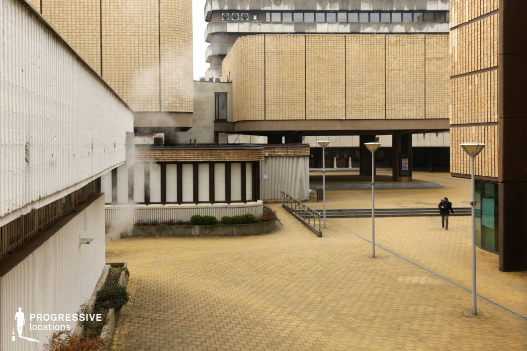 This expansive courtyard is surrounded by large brutalist-style buildings with textured yellow brick facades and stark concrete structures. The rigid geometric design, elevated walkways, and angular overhangs contribute to an imposing and institutional atmosphere. Tall, minimalistic street lamps line the open space, reinforcing the structured aesthetic. The combination of industrial elements and repetitive architectural patterns makes this an ideal location for dystopian, political thriller, or retro-futuristic film settings, evoking themes of surveillance, bureaucracy, or urban alienation.