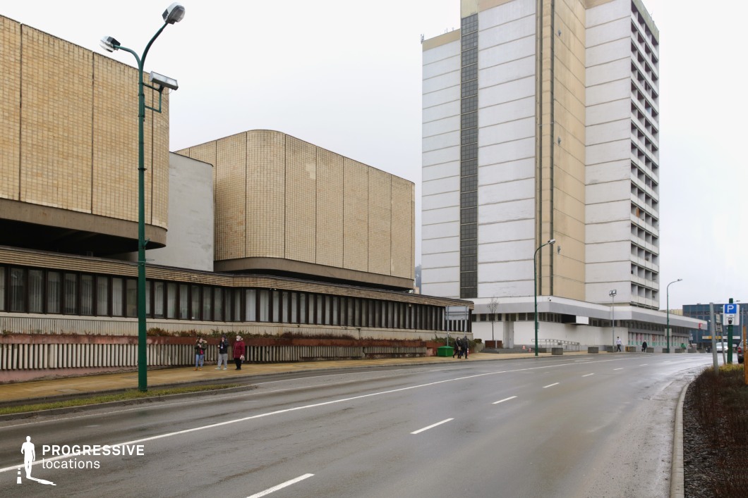 This urban setting features a striking example of brutalist architecture, with large, block-like buildings clad in beige tiles and concrete, emphasizing functionality over ornamentation. The combination of a towering high-rise and a lower, horizontally stretched structure creates a stark, institutional atmosphere, reinforced by the muted color palette and rigid geometric forms. The wide, multi-lane road adds a sense of openness, while the sparse placement of streetlights and minimal decorative elements contribute to the austere aesthetic. This location is ideal for filming dystopian, espionage, or neo-noir scenes, offering a cold, bureaucratic backdrop that evokes themes of surveillance, isolation, or state power. The imposing architecture also makes it suitable for historical or retro-futuristic settings, reminiscent of mid-to-late 20th-century socialist-era urban design.