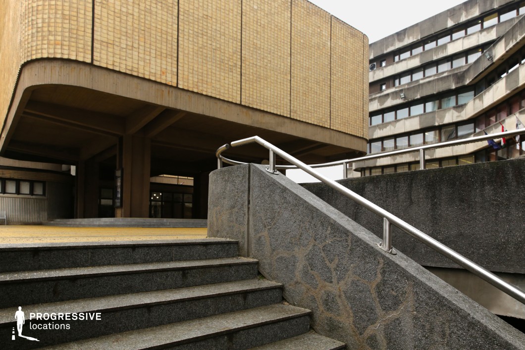 This imposing brutalist structure features raw concrete surfaces, angular geometric designs, and modular construction, embodying the stark, functional aesthetic of mid-to-late 20th-century socialist architecture. The elevated section, clad in uniform beige tiles, is supported by thick, heavy pillars, creating an open yet sheltered ground-level space. A wide concrete staircase with a minimalist metal railing leads toward the main entrance, adding to the monumental feel of the environment. In the background, a similarly designed weathered concrete building with ribbon windows reinforces the institutional character of the location. This setting is ideal for filming dystopian, political thriller, or cyberpunk narratives, offering a visually striking, rigid urban backdrop for tense confrontations, espionage sequences, or introspective character moments.