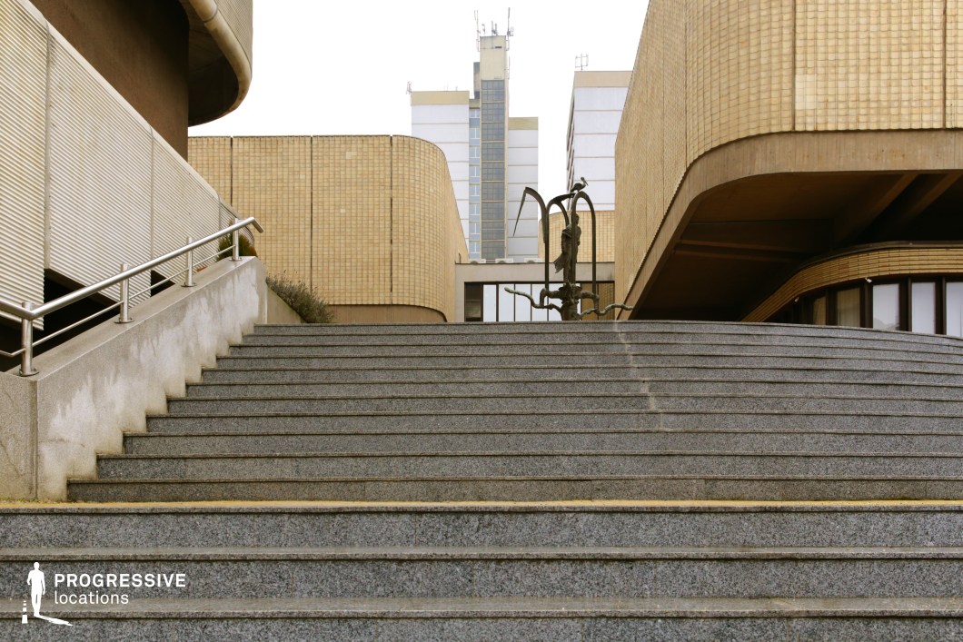 This image presents a striking composition of large, textured concrete steps leading towards a modernist architectural complex. The surrounding buildings, clad in yellowish brick and featuring curved and linear elements, exhibit a Brutalist and late-modernist design aesthetic. A sculptural metal installation at the top of the stairs adds an artistic focal point, while the symmetrical framing and geometric lines create a structured and imposing atmosphere. The mix of hard surfaces, sharp angles, and muted tones makes this location ideal for filming dystopian, political thriller, or corporate drama scenes, evoking themes of authority, power, and urban alienation.