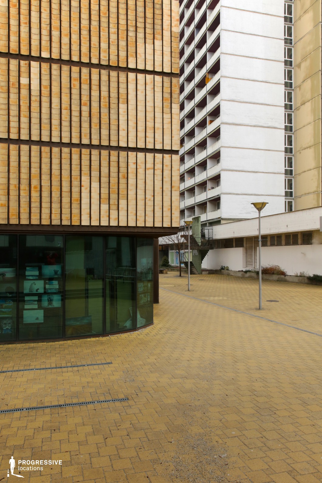 The image presents an urban courtyard framed by a mix of architectural styles. In the foreground, a modern curved structure features a glass facade and a textured, ochre-tiled exterior, creating a warm yet rigid geometric effect. Adjacent to it, a tall, utilitarian high-rise with repetitive balconies introduces a stark contrast, reinforcing the socialist-era architectural aesthetic. The courtyard is paved with yellow-toned bricks, extending an open, somewhat desolate atmosphere. Minimalist lampposts line the area, while an external staircase in the background adds depth and directionality to the scene. This setting is ideal for filming contemporary urban dramas, dystopian narratives, or social commentary pieces, offering a structured yet subtly oppressive environment suited for themes of isolation, transition, or nostalgia.