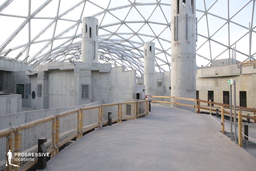 The image showcases a large, enclosed architectural space with a striking geodesic glass-and-metal dome overhead, allowing natural light to flood the area. The interior consists of raw concrete buildings and cylindrical towers with small, slotted openings, evoking an unfinished or industrial aesthetic. The elevated walkway, lined with bamboo and rope railings, curves through the structure, adding a contrast between natural and industrial elements. The muted gray tones of the concrete emphasize a utilitarian, dystopian atmosphere. This location is highly adaptable for science fiction, dystopian, or futuristic film settings, particularly for scenes involving research facilities, underground bunkers, or off-world colonies. The dramatic geometric ceiling structure enhances visual depth, making it an ideal setting for wide-angle cinematography and immersive storytelling.
