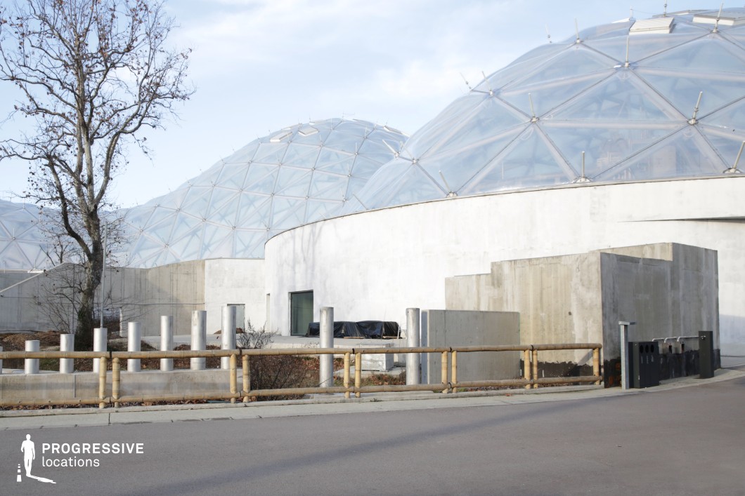 This location features a striking combination of geometric glass domes and raw concrete structures, creating a futuristic and industrial atmosphere. The domes are constructed with a transparent triangular framework, allowing natural light to filter through, while the surrounding buildings maintain a minimalist, utilitarian aesthetic with exposed concrete walls and angular formations. The juxtaposition of the curved domes with the rigid, box-like concrete sections adds visual contrast and depth. This setting is ideal for science fiction, dystopian, or high-tech thriller films, serving as a research facility, advanced greenhouse, or secret government installation. The open layout and structured design make it versatile for both wide establishing shots and intimate, tense close-ups.