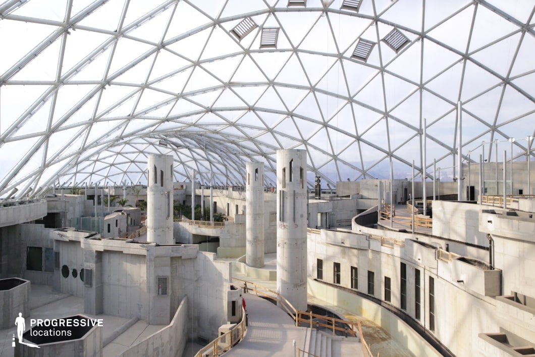 This expansive architectural space features a skeletal geodesic dome arching over an unfinished concrete structure with cylindrical towers and interconnected walkways. The raw concrete surfaces, sharp angles, and industrial aesthetics create a dystopian or futuristic atmosphere, ideal for science fiction, post-apocalyptic, or cyberpunk settings. The high ceiling and glass framework allow for dramatic lighting effects, making it suitable for visually striking action sequences, chase scenes, or psychological thrillers set in an enclosed yet vast environment.