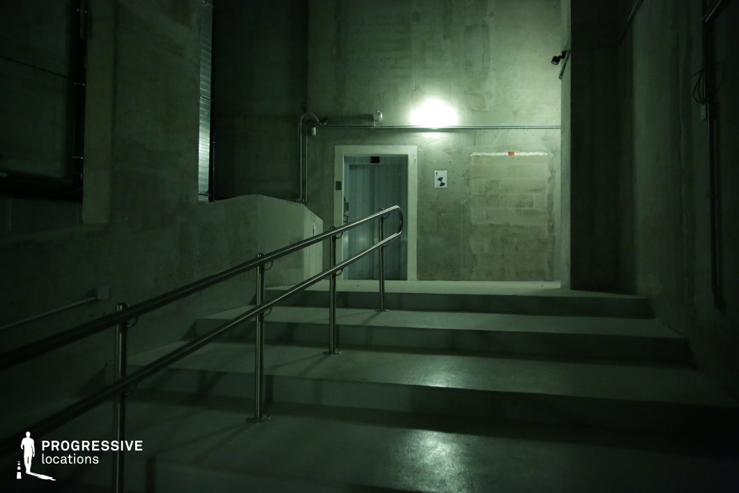 A dimly lit, industrial-style concrete stairwell with a metal handrail, surrounded by raw, unfinished walls. The surfaces are predominantly gray, with a smooth yet slightly worn texture, creating a stark, utilitarian aesthetic. The overhead fluorescent lighting casts a cold, greenish hue, enhancing the eerie and sterile ambiance. Exposed pipes and wiring contribute to the industrial, possibly institutional or underground setting. The closed metal door at the top of the stairs adds a sense of mystery, making the space ideal for suspenseful, dystopian, or crime thriller scenes. This location is well-suited for film sequences involving secrecy, tension, or restricted areas, fitting for genres such as horror, sci-fi, or espionage thrillers.