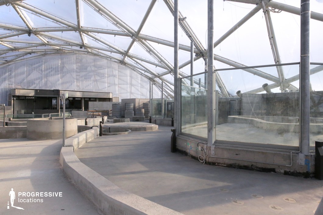 This location features a vast, semi-transparent glass and metal structure with a geometric framework, creating a futuristic, industrial aesthetic. The exposed metal beams crisscross overhead, forming an intricate lattice that enhances the visual depth of the space. Concrete pathways and barriers define the layout, contributing to a raw, unfinished look. The surrounding glass walls and panels introduce reflections and distortions, adding a dynamic visual element. The open, spacious design and muted color palette of grays and silvers emphasize a cold, modern atmosphere. Ideal for filming sci-fi, dystopian, or high-tech industrial settings, this space can serve as a research facility, secret base, or futuristic urban environment.
