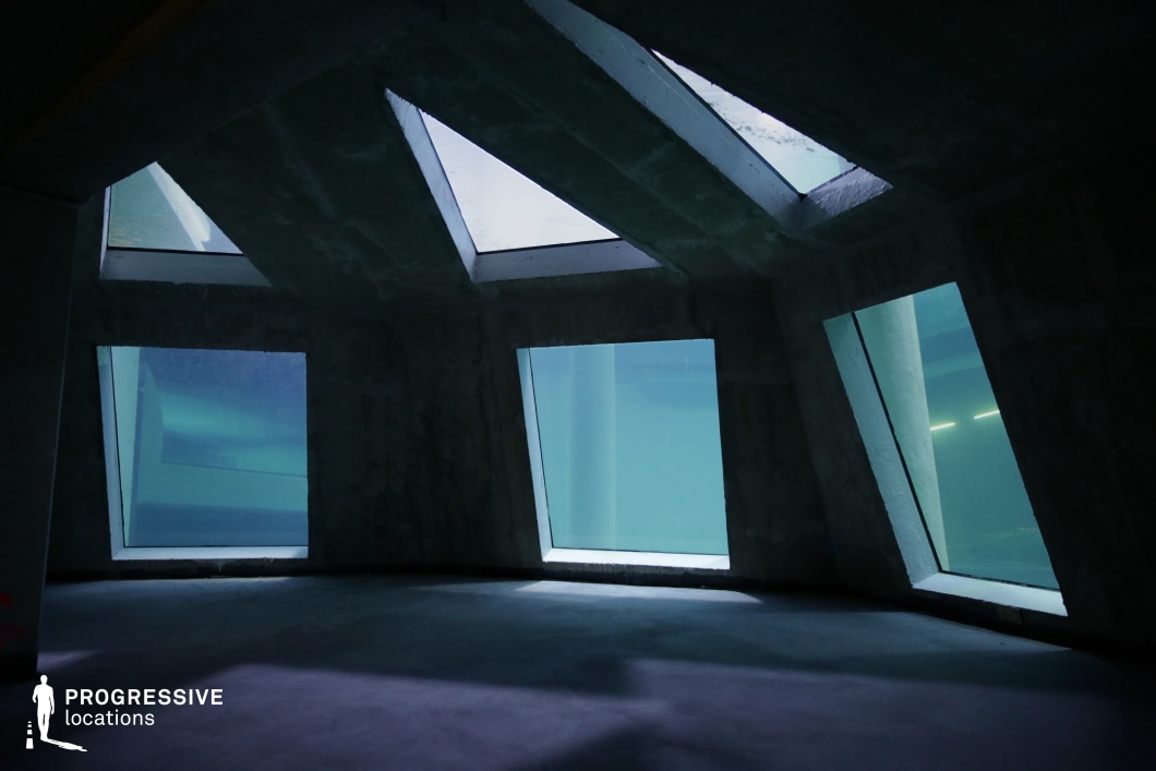 This unique space features an angular concrete interior with large, slanted glass panels offering a striking underwater view. The geometric windows, framed by rough-textured concrete, create a futuristic and industrial aesthetic, while the blue-green hues of the surrounding water add an atmospheric, almost otherworldly quality. The absence of furnishings and the stark contrast between light filtering through the water and the dark shadows on the floor enhance its cinematic potential. This location is ideal for sci-fi, mystery, or psychological thriller scenes, serving as an underwater laboratory, secret facility, or high-tech bunker.