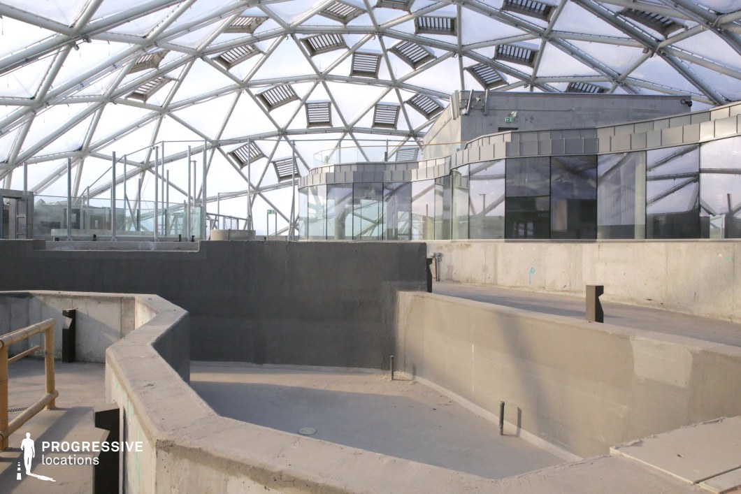 This location features a striking blend of industrial and modern architectural elements, dominated by raw concrete surfaces, geometric glass walls, and a transparent domed ceiling supported by an intricate metal framework. The open space, characterized by its angular, multi-level concrete partitions, creates a structured yet unfinished atmosphere. The reflective glass panels contrast with the rawness of the concrete, enhancing the futuristic and high-tech aesthetic. The geometric ceiling structure allows diffused natural light to filter through, adding depth and dimension to the space. This location is ideal for sci-fi, dystopian, or high-tech corporate thriller scenes, offering a visually compelling and versatile backdrop for futuristic settings or experimental architectural cinematography.