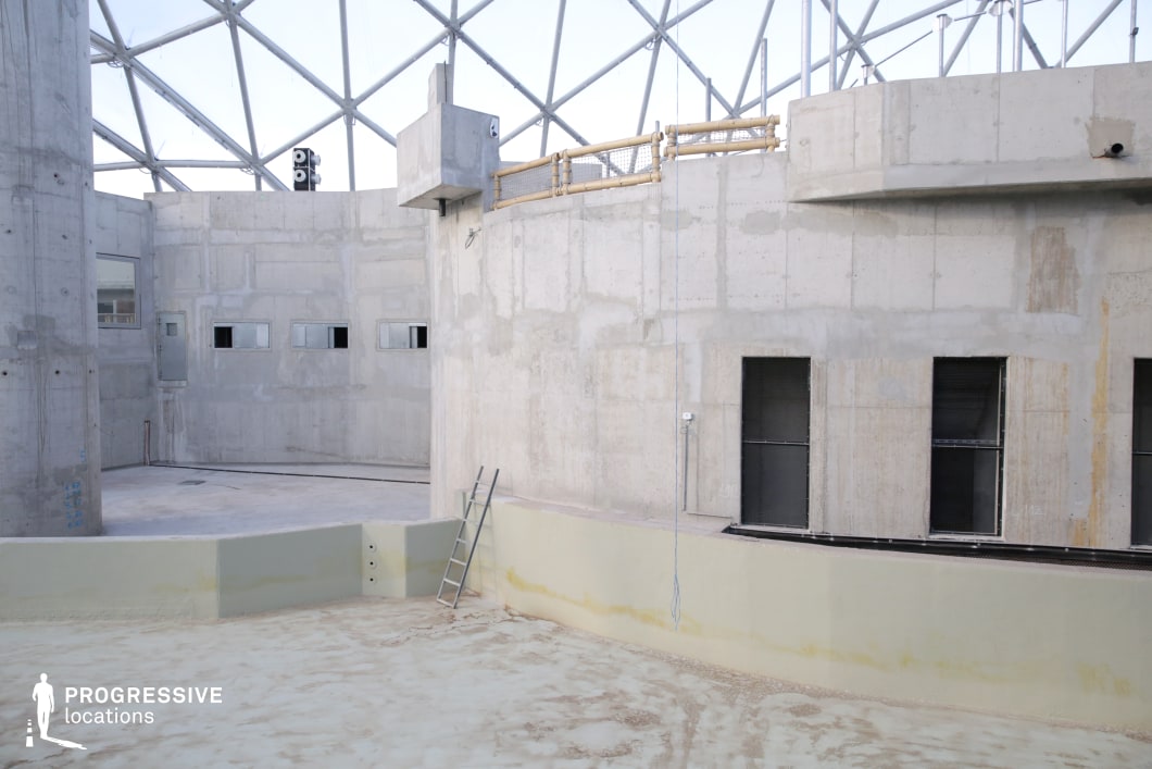 The image showcases a raw, unfinished concrete structure enclosed by a geodesic dome framework, creating a stark, industrial aesthetic. The walls are bare and textured, with small, rectangular windows and large open doorways adding an imposing, almost institutional feel. A metal ladder leans against a raised platform, suggesting an abandoned or repurposed space. The geometric metal framework above introduces a futuristic element, contrasting with the rough concrete surfaces. This location is ideal for dystopian, sci-fi, or industrial-themed film scenes, serving as a setting for secret facilities, research labs, or underground bunkers. The vast, open composition and layered architecture provide dramatic angles for cinematography.