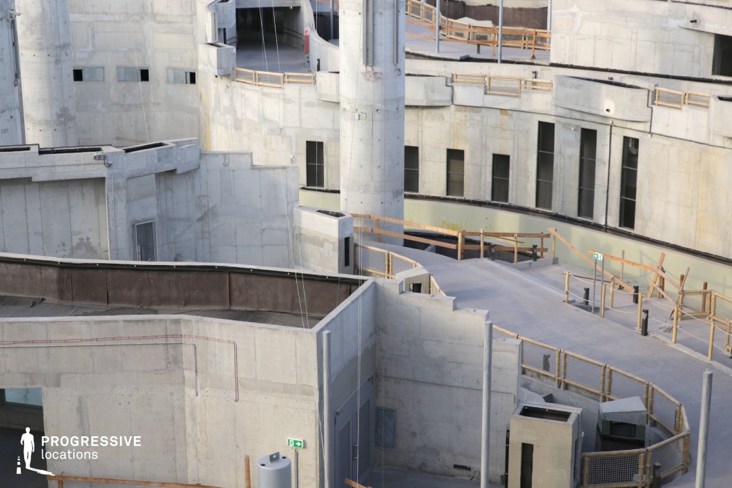 This location features an expansive, multi-tiered brutalist concrete structure with interconnected ramps, staircases, and elevated walkways. The raw, unfinished concrete surfaces and geometric design create an industrial, dystopian atmosphere. The space is characterized by angular shapes, rigid lines, and a stark color palette dominated by shades of gray. Wooden railings and metal mesh barriers contrast with the heavy concrete, adding a sense of temporary construction or decay. The setting is ideal for filming futuristic, sci-fi, or post-apocalyptic scenes, as well as action sequences requiring vertical movement and dynamic camera angles. Its labyrinthine layout also makes it suitable for chase scenes, psychological thrillers, or urban exploration narratives.