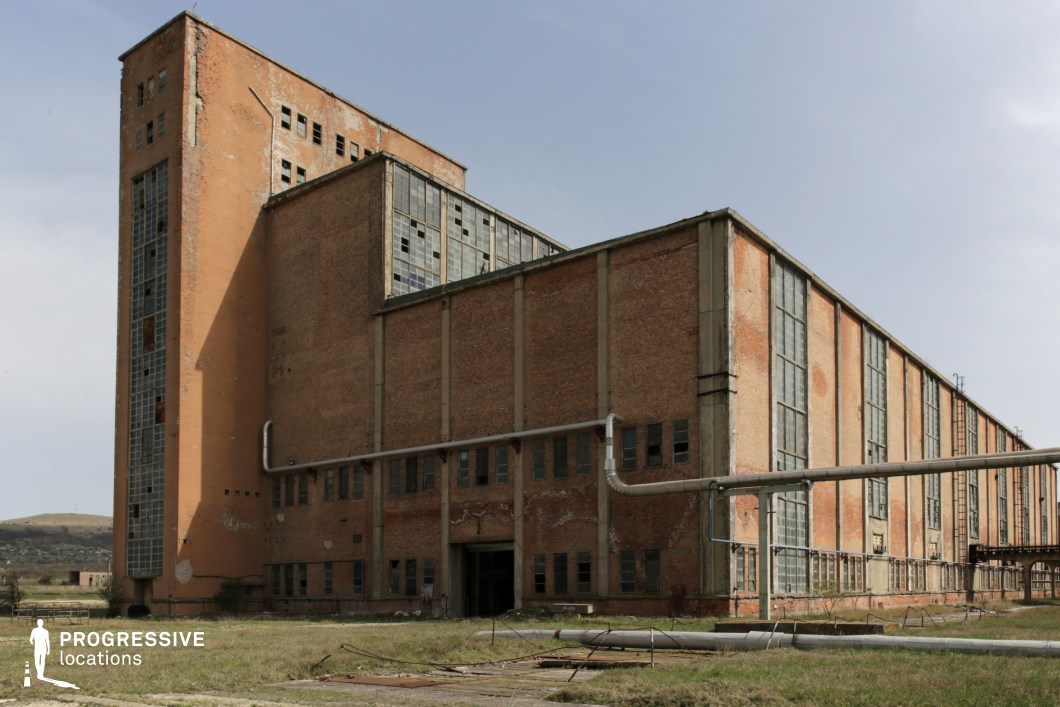 This expansive industrial complex, constructed primarily from weathered red brick, exudes a sense of decay and abandonment. The towering vertical section, with large multi-pane windows—many of them broken—adds a monolithic, almost oppressive feel to the site. The long horizontal structure, reinforced by concrete pillars, features rusting metal pipes running along its facade, further emphasizing its former industrial function. Overgrown grass and scattered debris in the foreground enhance the location’s post-industrial aesthetic. This setting is ideal for filming dystopian, war, or crime-themed productions, providing an imposing backdrop for scenes involving urban decay, abandoned infrastructure, or clandestine operations. The combination of industrial scale and deteriorating architecture makes it a compelling choice for gritty, atmospheric storytelling.