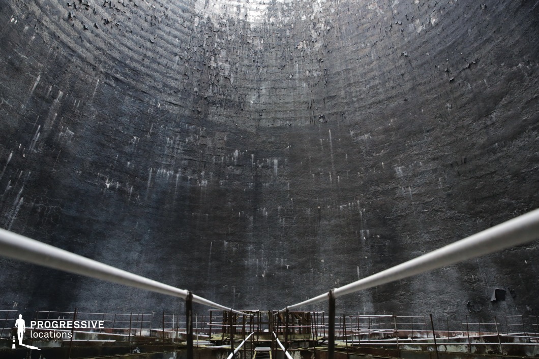 This massive circular structure features towering, weathered concrete walls with a rough, textured surface, creating an overwhelming sense of scale and isolation. The decayed interior is marked by peeling layers of dark material, streaked by water damage, giving it an eerie, post-apocalyptic atmosphere. A rusted metal walkway extends into the vast chamber, adding depth and perspective. The dramatic curvature of the walls enhances the acoustics and visual intensity of the space. This location is ideal for filming sci-fi, dystopian, or horror scenes, serving as an underground bunker, secret industrial site, or a desolate sci-fi environment. The enclosed, cavernous nature of the space allows for striking cinematographic compositions, playing with shadows and scale.