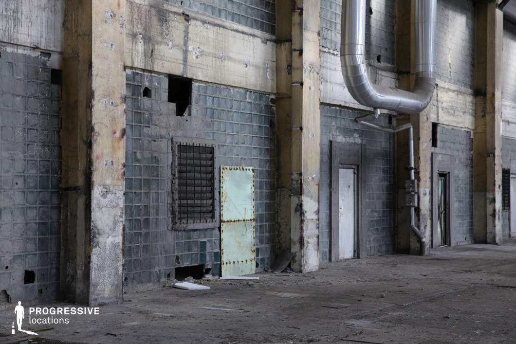 This deteriorating industrial space features a wall of aged glass bricks, corroded metal doors, and exposed, discolored concrete columns. The cracked surfaces and broken ventilation grates contribute to the building’s decayed and neglected atmosphere. Large, curved metal pipes run along the walls, hinting at the site's former mechanical or industrial function. The grimy floor and scattered debris enhance the sense of abandonment. This location is ideal for dystopian, post-apocalyptic, or crime-thriller settings, offering a gritty, raw aesthetic perfect for tense confrontations, hideouts, or clandestine operations. The industrial textures and muted color palette provide a striking cinematic backdrop.