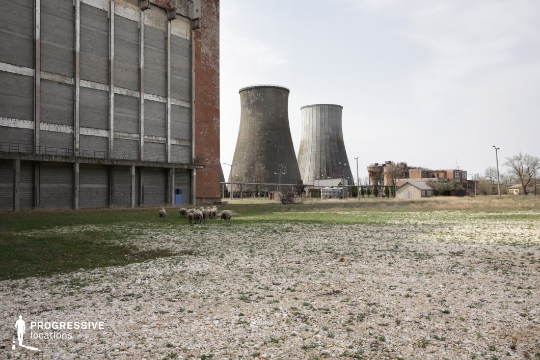 This expansive industrial landscape features massive cooling towers and a weathered brick-and-concrete structure, evoking a sense of abandonment and decay. The imposing, monolithic walls of the nearby building contrast with the open, gravel-covered foreground, creating a striking blend of industrial brutalism and desolation. The distant, rusting factory elements and small outbuildings contribute to the post-industrial atmosphere. This location is ideal for dystopian, sci-fi, or post-apocalyptic storytelling, offering a dramatic and eerie backdrop for sequences involving environmental collapse, corporate secrecy, or a world reclaimed by nature.