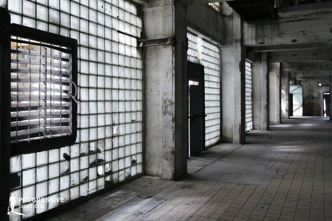 This desolate industrial corridor features decayed concrete walls, grid-patterned glass block windows, and a floor covered in worn, cracked tiles. The translucent glass bricks diffuse dim light, casting a moody glow that enhances the aged and abandoned feel of the space. A metal-barred vent disrupts the symmetry, while broken glass blocks add an element of neglect. The repetitive columns and doorways create a strong sense of depth and perspective, ideal for tense, suspenseful sequences. This location is well-suited for thriller, horror, or dystopian settings, providing a visually striking backdrop for escape, confrontation, or mystery-driven narratives.