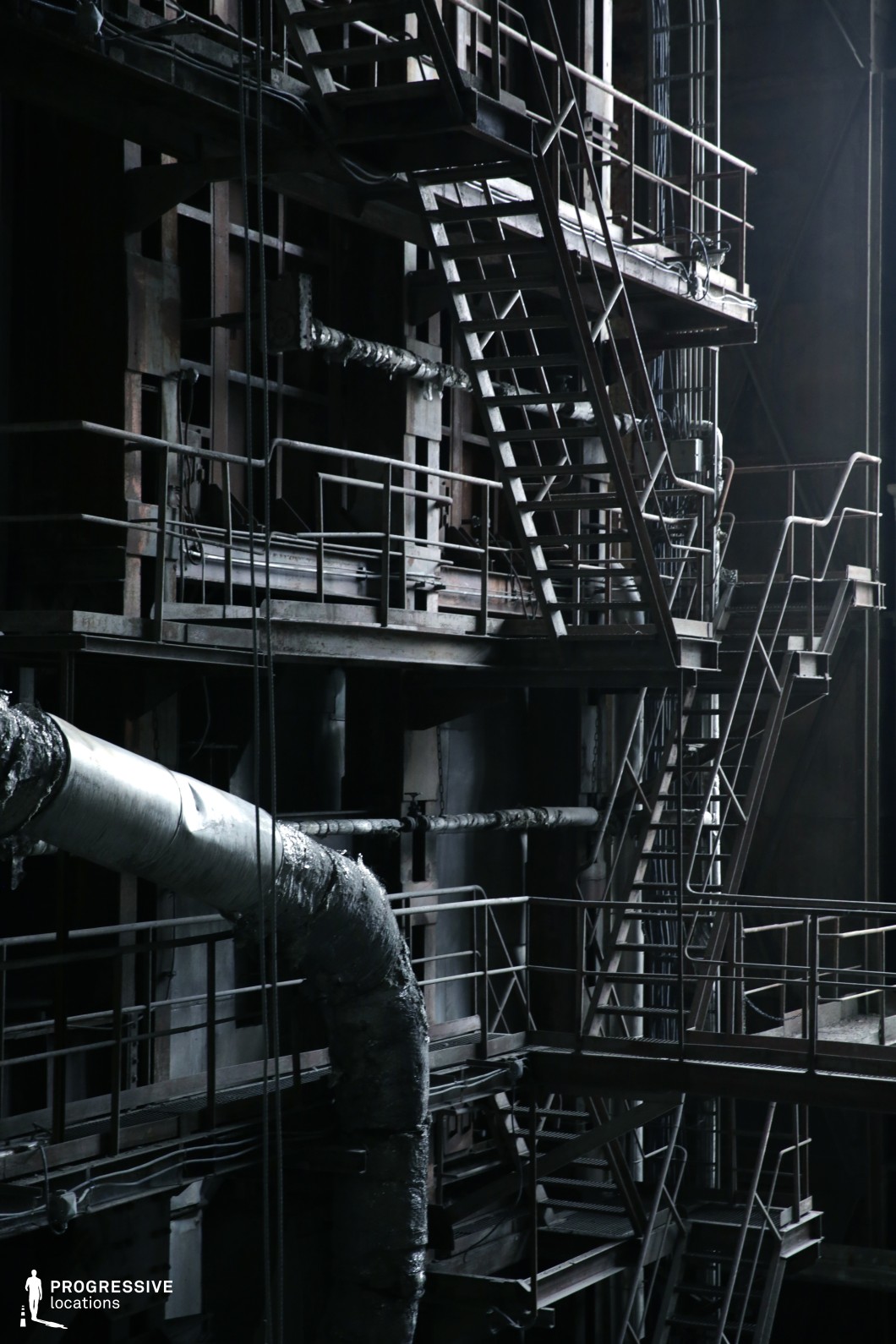 This intricate industrial environment is dominated by a network of rusted metal staircases, catwalks, and platforms, forming a labyrinthine structure of intersecting lines and geometric patterns. The dim lighting and deep shadows enhance the sense of mystery and danger, while exposed pipes and aging metal surfaces reinforce the setting’s industrial decay. The worn insulation on the large ventilation duct adds to the gritty, abandoned aesthetic. This location is ideal for cyberpunk, thriller, or action films, serving as a high-stakes chase scene, a clandestine meeting spot, or a secret underground facility. The complex interplay of shadows and structural elements allows for dynamic cinematography, emphasizing tension and vertical movement.