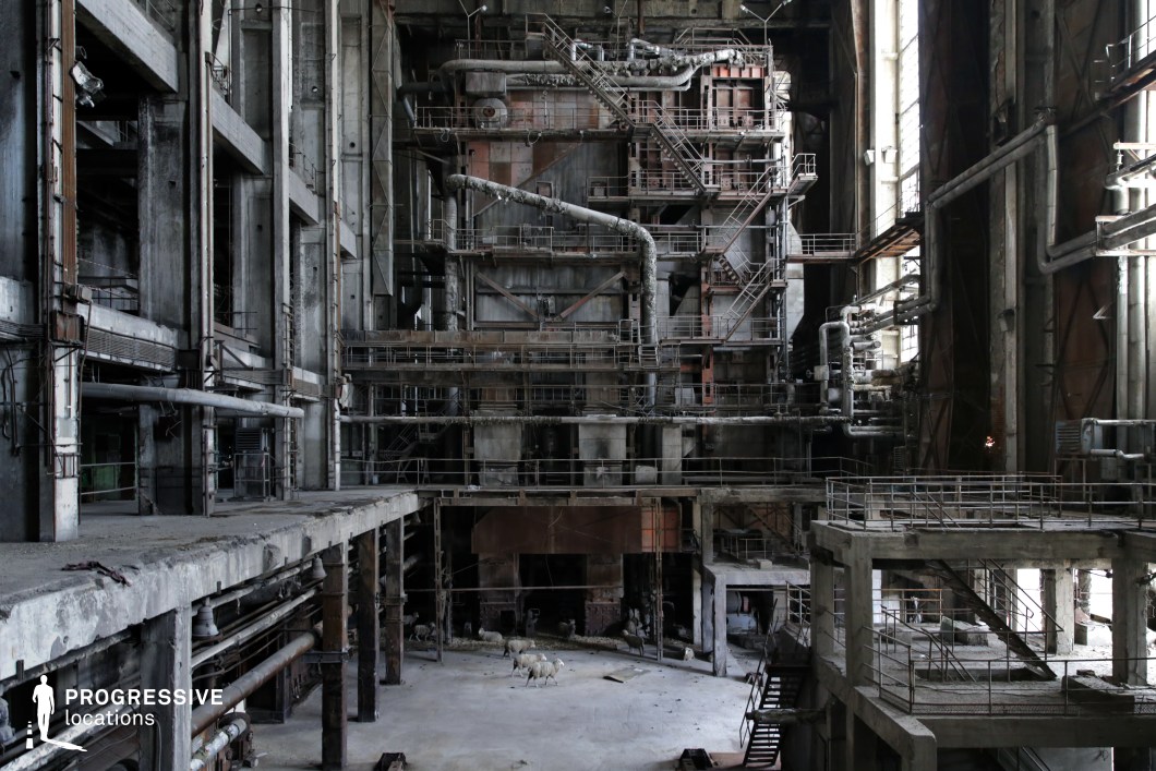 The image showcases a vast, abandoned industrial facility with towering metal structures, corroded pipes, and multiple levels of walkways interconnected by staircases. The exposed concrete walls and rusted steel elements create a stark, dystopian aesthetic, further emphasized by the dim lighting filtering through the high windows. The cavernous space, marked by decayed surfaces and scattered debris, suggests a former power plant or large-scale factory, now left to decay. This setting is ideal for filming post-apocalyptic, dystopian, cyberpunk, or thriller scenes, providing an immersive backdrop for abandoned world-building, intense chase sequences, or clandestine meetings in an industrial wasteland.