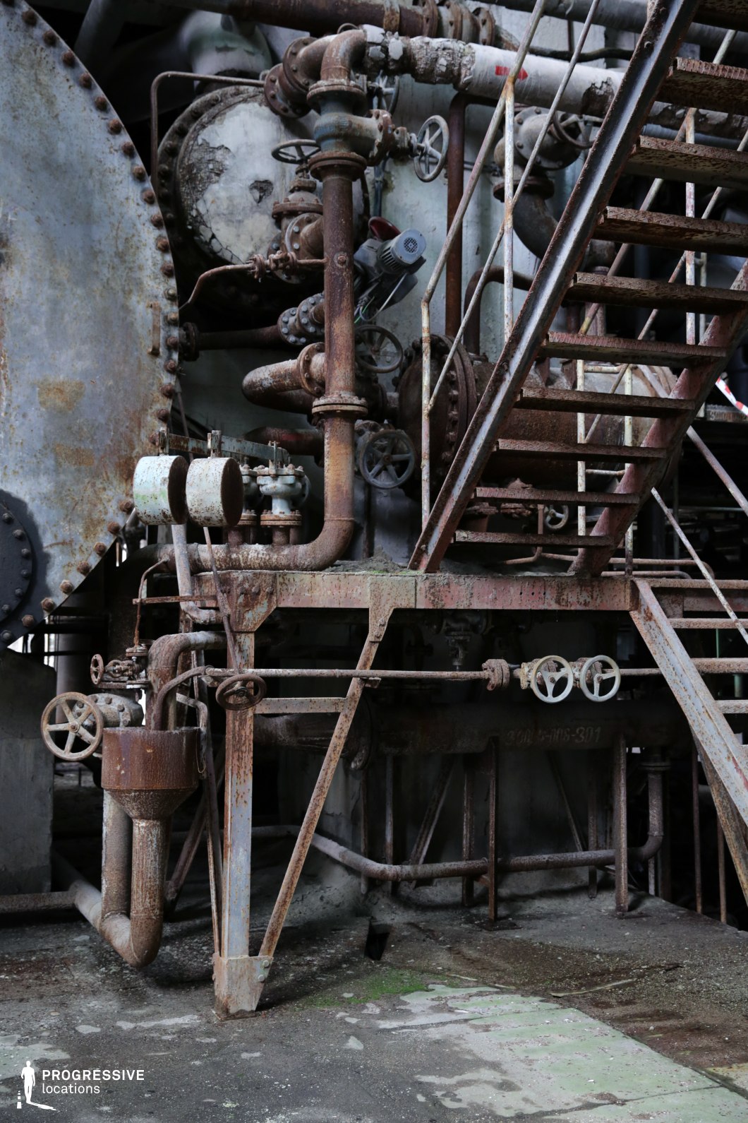 The image showcases an aged industrial setting dominated by corroded steel pipes, valves, and machinery, evoking a sense of abandonment and decay. The intricate network of interconnected pipes and rust-covered components suggests an old factory, power plant, or refinery. The dominant materials include heavily oxidized metal, contributing to a gritty, industrial aesthetic. A rusted metal staircase with worn steps ascends diagonally, adding depth and structural complexity to the scene. The muted color palette, consisting of browns, grays, and faded greens, enhances the raw and gritty atmosphere. This location is ideal for filming dystopian, post-apocalyptic, or industrial thriller scenes, providing an authentic setting for narratives involving decay, mystery, or clandestine operations.