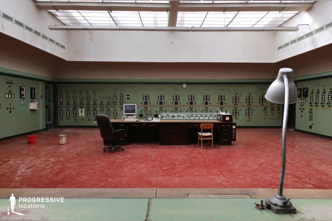 This abandoned industrial control room features a striking mid-century aesthetic with a green panel-lined wall filled with analog dials, switches, and meters. The red, worn-out flooring contrasts with the muted tones of the walls, reinforcing the aged, utilitarian atmosphere. A central wooden desk, cluttered with old office equipment, dominates the room, accompanied by mismatched chairs that enhance the retro, functionalist aesthetic. The ceiling, framed by a glass skylight, allows diffused natural light to illuminate the space, emphasizing the faded textures and peeling paint on the upper walls. This location is ideal for film productions requiring a vintage industrial setting, making it a prime choice for historical dramas, dystopian thrillers, or sci-fi narratives that demand a cold war-era or abandoned facility ambiance.