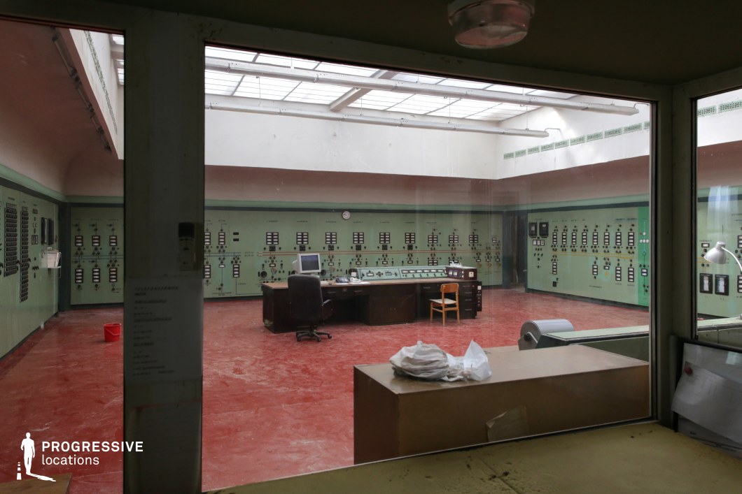 This industrial control room features an aged, utilitarian design with green-painted control panels lining the walls, filled with analog switches, dials, and meters, evoking a mid-20th-century industrial aesthetic. The floor is a striking red, contrasting with the muted green walls, while overhead skylights allow natural light to diffuse into the space. A central workstation with a dark wooden desk and outdated computer monitors sits at the heart of the room, reinforcing its vintage, possibly decommissioned, atmosphere. The worn-down condition, combined with the stark fluorescent lighting fixtures, enhances the space's eerie and utilitarian character. This setting is ideal for filming scenes that require a retro-futuristic, dystopian, or Cold War-era control center ambiance. It would be particularly effective for sci-fi, espionage, or thriller genres, portraying secrecy, decay, or technological antiquity.