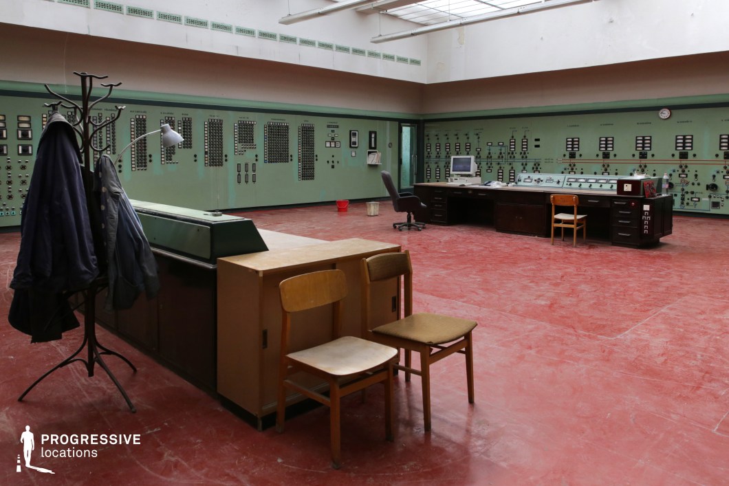 This industrial control room features an expansive layout with green control panels lining the walls, filled with dials, switches, and meters, evoking a mid-20th-century technological aesthetic. The worn red floor contrasts with the muted green tones, reinforcing a sense of aged functionality. Overhead skylights provide diffused natural light, highlighting the utilitarian design. A mix of wooden and metal furnishings, including desks, chairs, and a coat rack, contribute to the space’s retro-futuristic, slightly abandoned atmosphere. Ideal for filming period sci-fi, Cold War-era thrillers, or dystopian narratives, this location serves as a compelling backdrop for tension-filled or surveillance-heavy scenes.