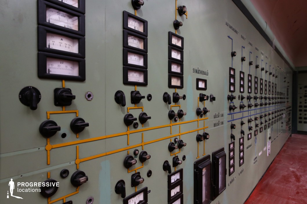 This vintage control panel features an extensive array of analog gauges, rotary switches, and intricate wiring diagrams set against a muted green metal surface. The symmetrical layout of dials and meters, with bold black casings and yellow and blue circuit lines, suggests a mid-20th-century industrial or power station setting. The aged paint and worn textures add to its retro, utilitarian aesthetic, evoking a time when manual control and monitoring were essential. The narrow, tunnel-like perspective created by the long panel enhances the sense of depth and immersion, making this an ideal setting for historical, dystopian, or sci-fi productions. Its distinctive look is well-suited for scenes requiring a Cold War-era atmosphere, government facility interiors, or secret underground control centers.