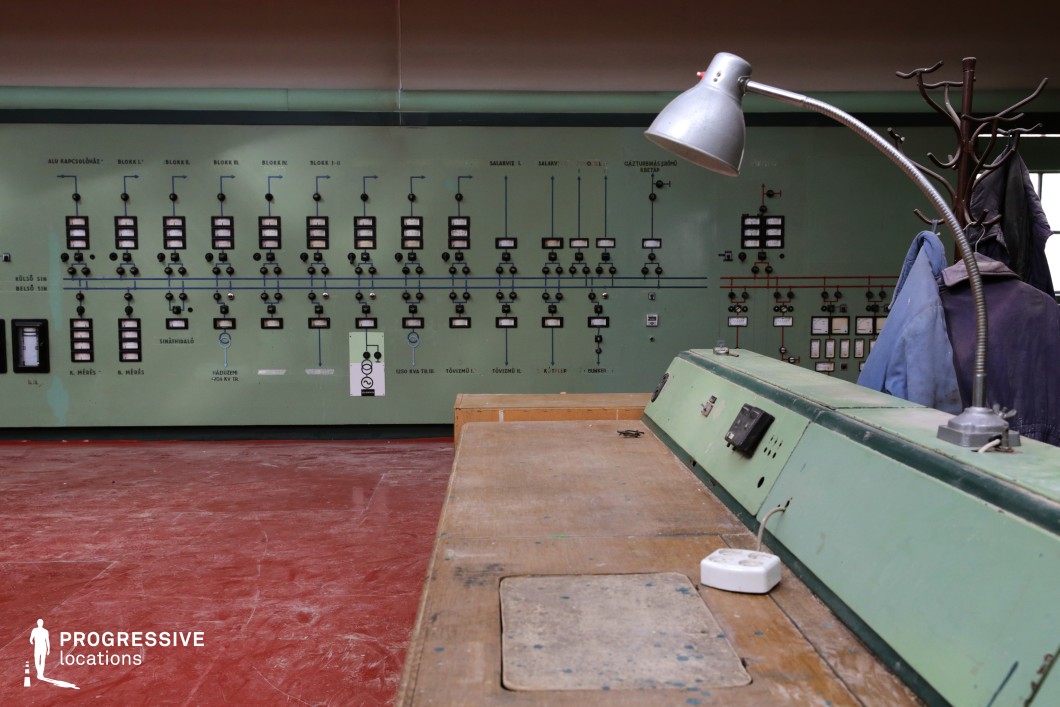 The image showcases an aged industrial control room with a distinctly retro aesthetic. The dominant green control panel spans the background, filled with an array of analog dials, switches, and meters, suggesting mid-to-late 20th-century technology. A worn wooden desk with a built-in green metal console is positioned in the foreground, adding to the workspace’s utilitarian feel. A flexible metallic desk lamp arches over the station, contributing to the old-school industrial atmosphere. The red flooring provides a striking contrast to the muted green tones, reinforcing the stark and functional design. The setting evokes a Cold War-era or Soviet-style industrial environment, making it ideal for filming historical, dystopian, or espionage-themed productions. This location is well-suited for scenes set in abandoned facilities, secretive government installations, or vintage technical operations.