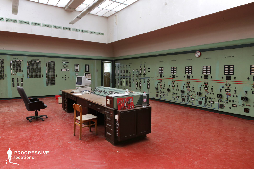 This industrial control room features a retro-futuristic aesthetic with muted green walls lined with an array of analog dials, switches, and control panels, evoking a mid-20th-century technological environment. A large wooden desk with an integrated control console sits at the center, surrounded by worn office chairs, hinting at a space once active but now frozen in time. The red linoleum flooring contrasts starkly with the pale green walls, reinforcing a utilitarian, almost socialist-realist atmosphere. Overhead, a skylight provides natural illumination, emphasizing the room’s aged yet functional appearance. The space is well-suited for film productions requiring an old-fashioned industrial or bureaucratic setting, making it ideal for science fiction, historical thrillers, Cold War-era dramas, or dystopian narratives.