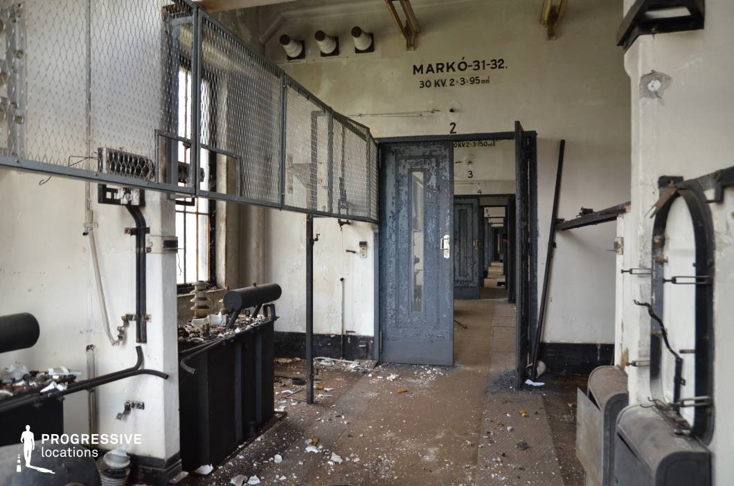 This decayed industrial facility features a long, narrow corridor lined with rusted doors, peeling paint, and exposed wiring, exuding an eerie, abandoned atmosphere. The high walls are marked with technical inscriptions, suggesting a former electrical or mechanical use, while broken fixtures and scattered debris add to the sense of dereliction. Overhead metal mesh barriers and corroded pipes reinforce the utilitarian, harsh aesthetic. The space is ideal for filming tense, dystopian, or post-apocalyptic scenes, as well as crime, horror, or investigative thriller narratives requiring an ominous, forsaken industrial backdrop.