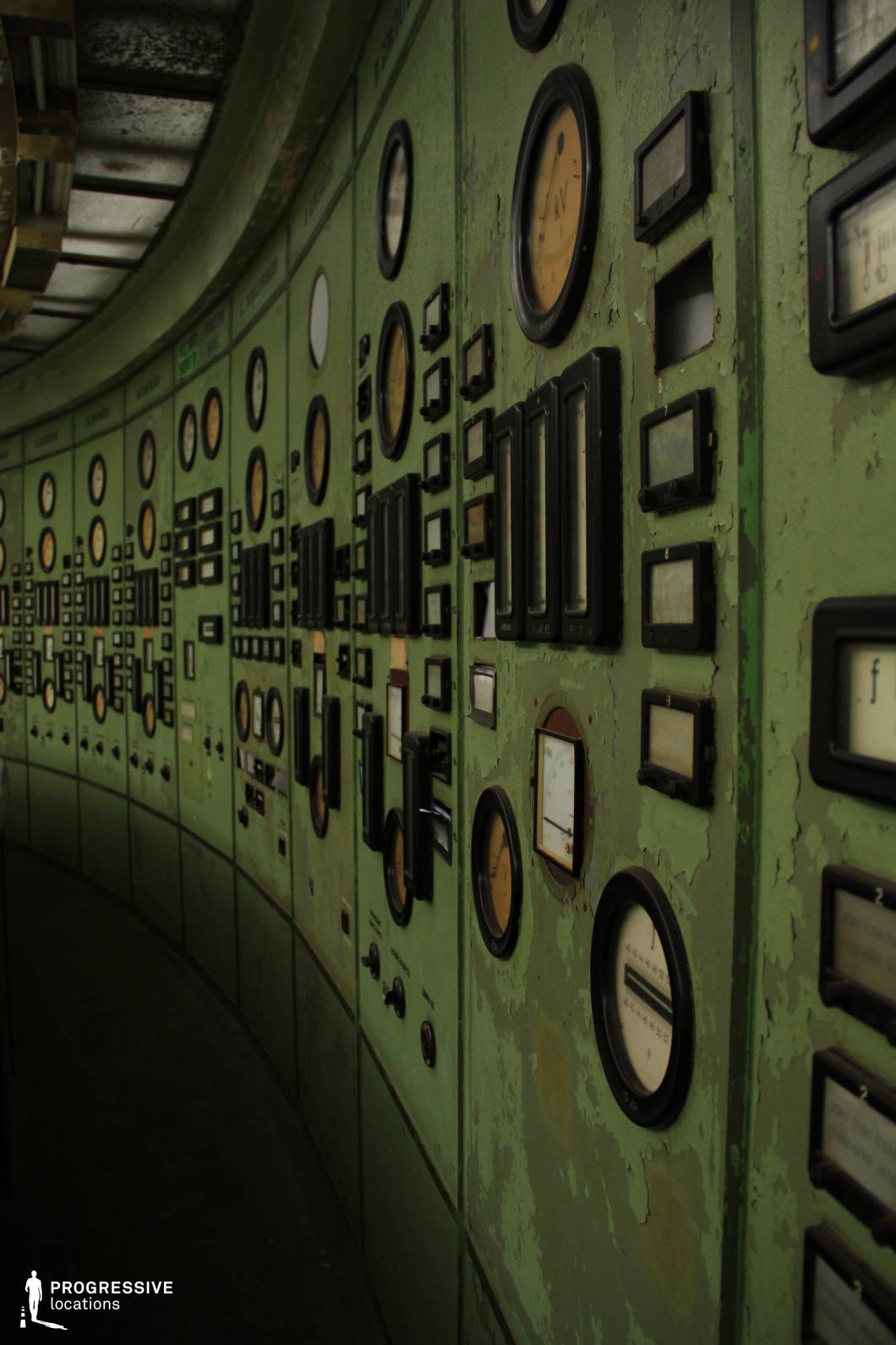 This long, curved industrial control panel is covered in an array of analog gauges, switches, and meters, all set against a faded green metal surface with peeling paint and visible wear. The dim lighting and the heavy, mechanical aesthetic of the panel suggest a historical industrial or power facility, evoking a sense of outdated technology and abandonment. The intricate details of dials and switches create a visually rich, textured scene with a strong retro-futuristic or Cold War-era atmosphere. This location is ideal for filming scenes set in a dystopian, sci-fi, or thriller context, offering a setting that conveys secrecy, decay, or the remnants of a once-powerful technological infrastructure.
