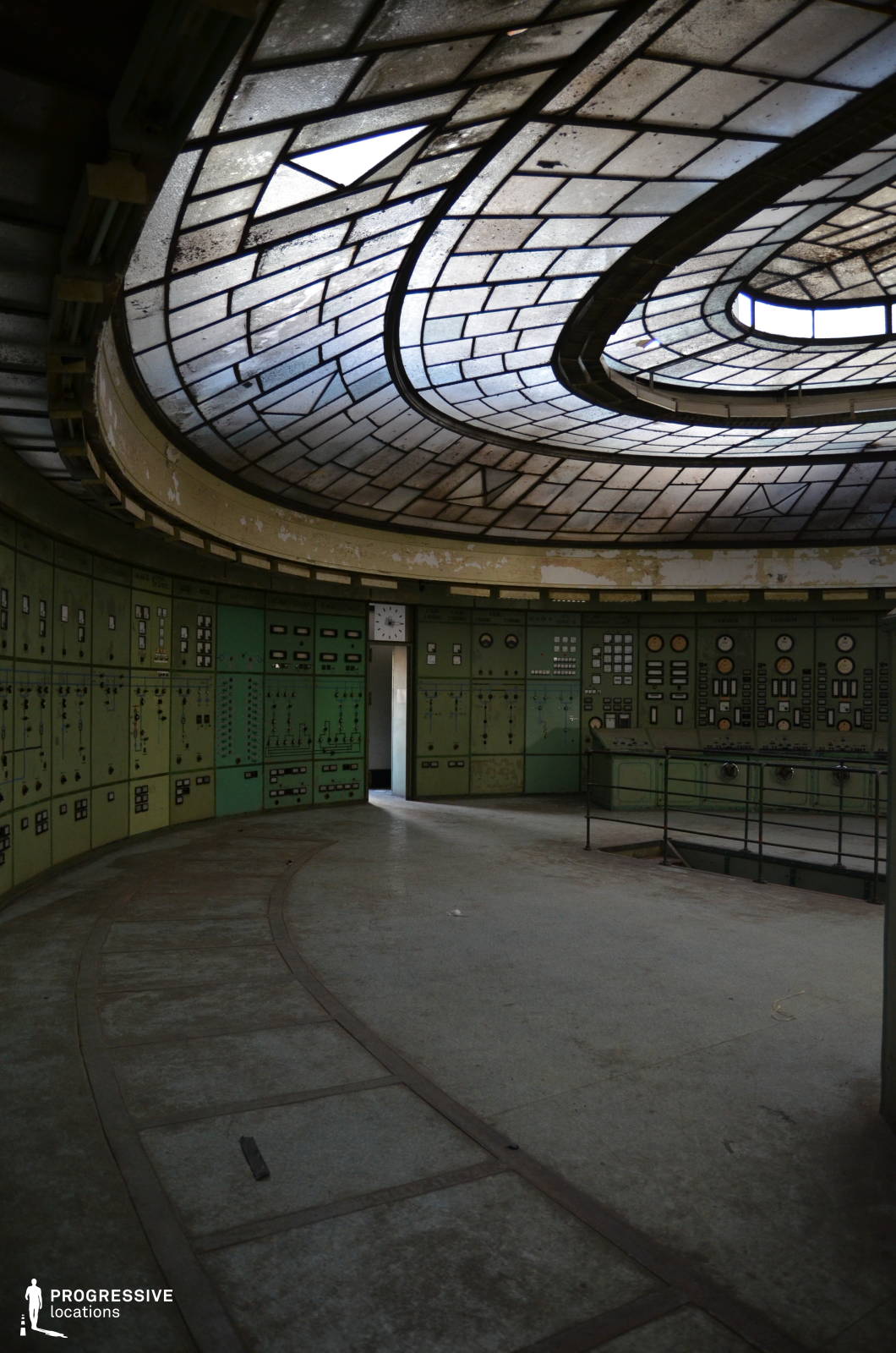 This circular industrial control room features a dramatic curved glass ceiling, composed of aged, geometric panels that filter dim natural light into the space. The walls are lined with vintage green control panels, densely packed with analog dials, switches, and gauges, reinforcing the mid-century industrial aesthetic. The peeling paint and worn flooring add to the room’s abandoned, timeworn character, while the curved layout enhances the immersive, enclosed feel of the environment. The setting is perfect for sci-fi, dystopian, or historical thrillers, offering an ideal backdrop for scenes depicting secrecy, technological decay, or authoritarian control.
