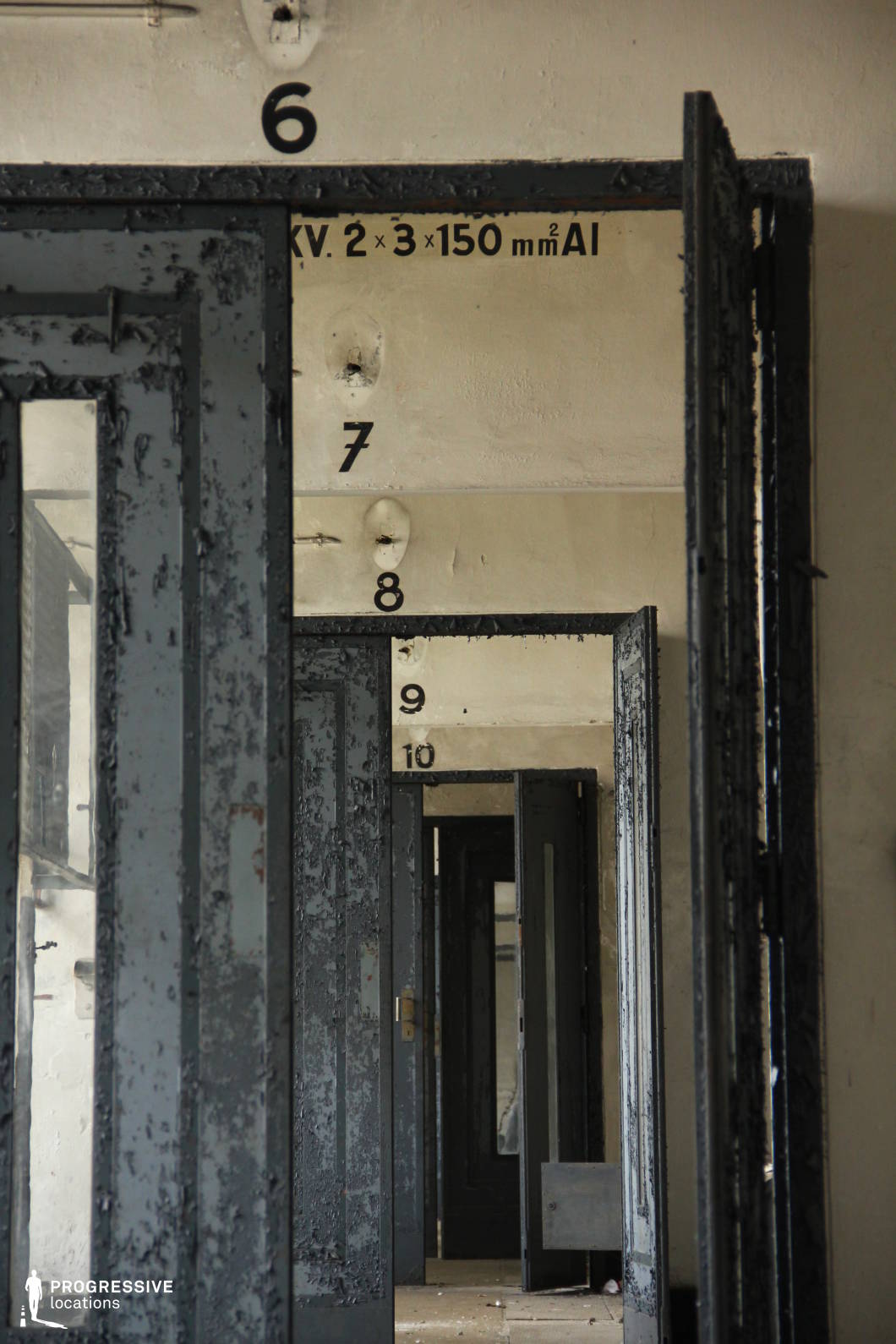 The image showcases a long, narrow corridor lined with heavy, industrial-style metal doors that are peeling and rusted, evoking a sense of abandonment. The sequence of open doors creates a strong visual depth, emphasizing repetition and perspective. The walls, marked with numbering and technical annotations, suggest a utilitarian or institutional function, possibly an old prison, military facility, or industrial site. The muted, neutral color palette of gray and beige, combined with the worn textures of metal and concrete, establishes a stark and oppressive atmosphere. This location is ideal for filming psychological thrillers, horror, dystopian settings, or any scene requiring a tense, confined environment with a sense of decay and history.