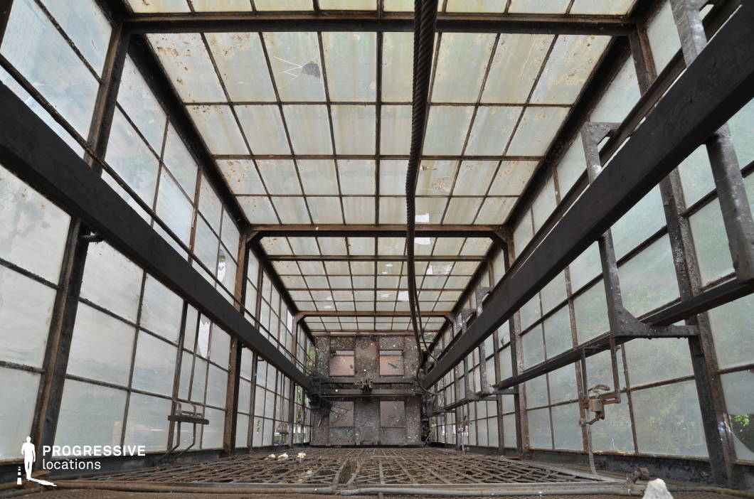 This towering industrial elevator shaft, enclosed by rusted metal frames and translucent glass panels, exudes a sense of decay and abandonment. The glass, now weathered and cracked in places, allows diffused natural light to filter through, creating an eerie, muted glow that enhances the aged aesthetic. The exposed metal beams and gridded flooring contribute to the harsh, utilitarian design, while the visible wear, rust, and dirt add a layer of historical depth. The vertical perspective emphasizes height and depth, making this location visually striking. This setting is ideal for filming thriller, dystopian, or post-apocalyptic scenes, evoking isolation, tension, or the remnants of forgotten industry. The structural elements also provide dynamic opportunities for cinematography, especially for suspenseful or escape-oriented sequences.