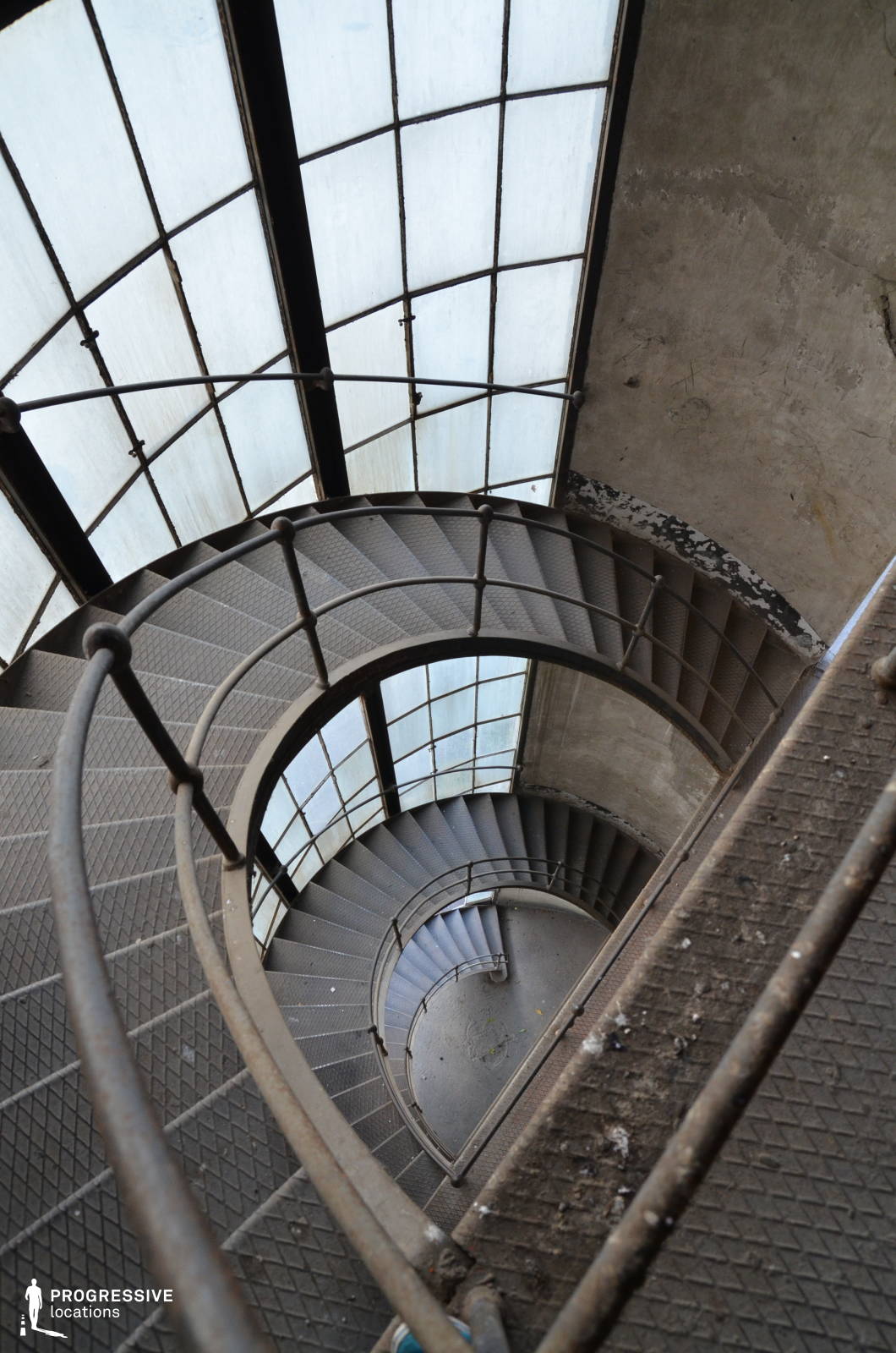 This striking spiral staircase features textured metal steps and a weathered handrail, winding downward in a mesmerizing geometric pattern. The surrounding industrial-style glass windows, divided by aged metal frames, filter soft, diffused light into the space, contrasting with the raw, unfinished concrete walls. The chipped paint and worn surfaces enhance the aged, utilitarian character of the setting. The combination of curved lines and rigid structural elements creates a dynamic visual composition, making this location ideal for psychological thrillers, noir-inspired cinematography, or historical dramas. The staircase’s dramatic perspective lends itself to chase sequences, moments of introspection, or mysterious, suspenseful storytelling.