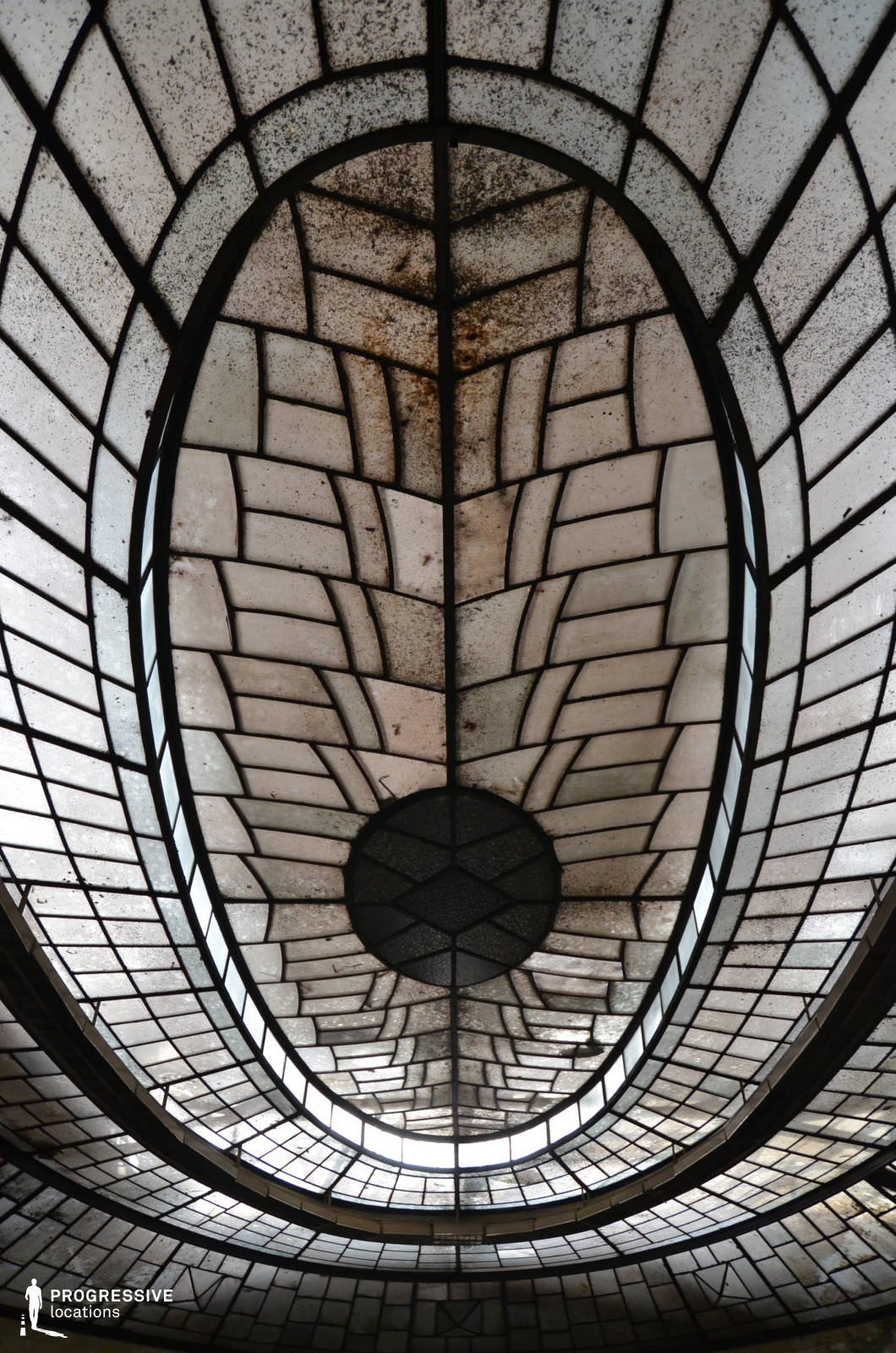 This striking oval-shaped glass ceiling features an intricate geometric pattern, reminiscent of Art Deco or early modernist design. The segmented glass panels, framed by dark metal, create a structured yet organic flow, converging at a circular centerpiece. The aged and slightly weathered appearance of the glass adds a historical character, suggesting an old industrial or civic building. The interplay of light filtering through the translucent panes enhances the dramatic atmosphere, making this location ideal for filming mysterious, historical, or futuristic settings. Perfect for noir thrillers, period dramas, or visually rich dystopian narratives.
