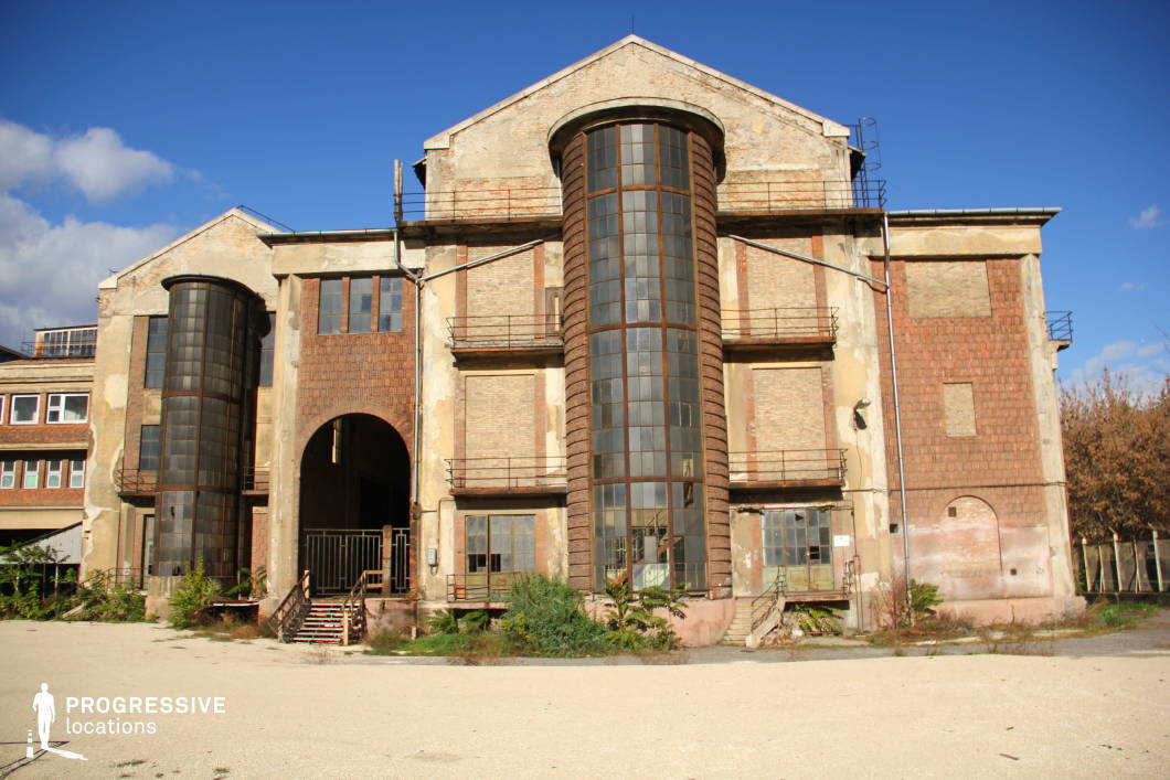 This imposing, decayed industrial building features a symmetrical façade with two large cylindrical glass towers flanking a central arched entrance. The exterior is a mix of exposed brick, faded plaster, and rusted metal elements, giving it a raw, weathered aesthetic. The metal balconies and staircases add an industrial-era charm, while the large, empty windows and peeling surfaces create a sense of abandonment. The surrounding open courtyard provides ample space for dynamic camera movement. This location is ideal for filming dystopian, post-apocalyptic, or historical narratives, as well as scenes depicting abandoned research facilities, secretive institutions, or forgotten power plants.