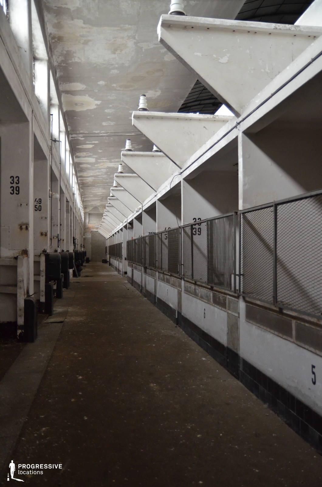 The image showcases a long, decayed corridor in what appears to be an old industrial or agricultural facility. The space is characterized by a repetitive series of numbered stalls separated by metal mesh fencing and reinforced concrete beams that form striking angular structures. The peeling paint on the ceiling and walls, along with the worn concrete floor, conveys a sense of abandonment and past utility. The perspective of the image, with its vanishing-point composition, creates a strong depth effect, making it an ideal location for filming tense, atmospheric, or dystopian scenes. This setting is well-suited for thriller, horror, or post-apocalyptic genres, as well as documentary-style storytelling about industrial decline.