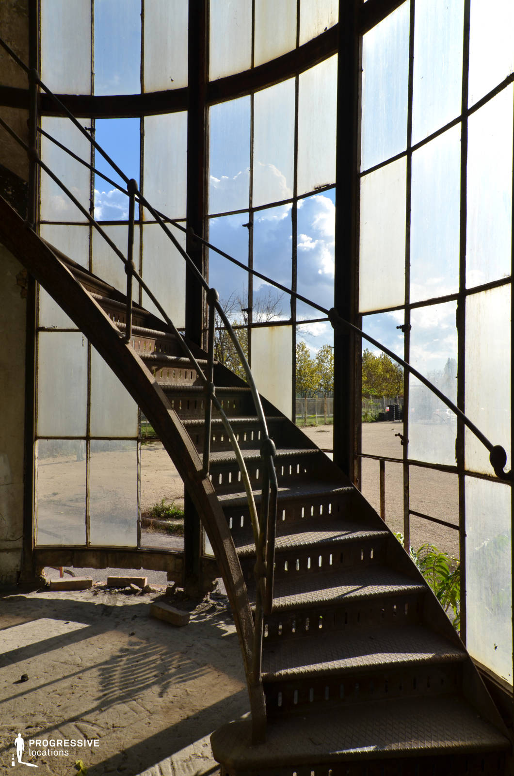 The image features a striking curved iron staircase within a semi-circular glass-walled structure, creating a dramatic blend of industrial and architectural aesthetics. The metal steps are perforated and aged, showing signs of wear, while the delicate railings curve in harmony with the overall structure. The large, segmented glass panes, some of which are broken or stained, provide a diffused and moody light, emphasizing the raw, abandoned atmosphere. The floor is covered in dust and debris, adding to the sense of decay. This location is ideal for filming atmospheric scenes in genres such as neo-noir, post-apocalyptic, or historical drama, lending itself well to mysterious, introspective, or suspenseful storytelling. The contrast between the decayed interior and the open outdoor space beyond the glass adds depth and visual intrigue.