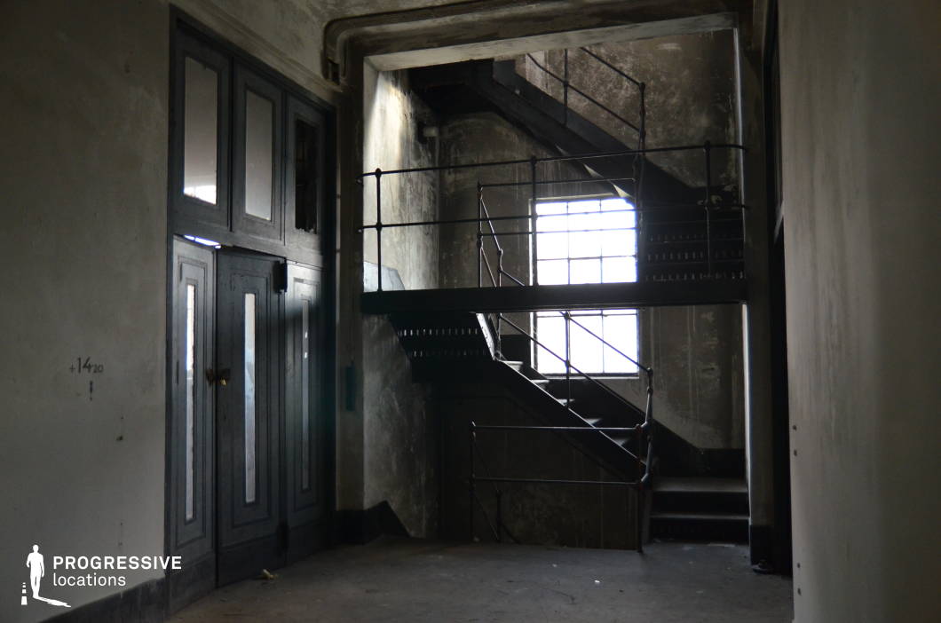 This dimly lit industrial stairwell exudes a raw, utilitarian aesthetic with its aged metal railings, dark wooden steps, and worn concrete walls. A large window casts diffused light into the space, creating stark contrasts between illuminated and shadowed areas. The heavy double doors with reflective glass panels add an element of mystery, while the peeling surfaces and exposed pipes suggest an abandoned or long-unused facility. The setting is ideal for filming tense, suspenseful scenes in thriller, noir, or dystopian genres, serving as an escape route, a confrontation zone, or an atmospheric transition space in a larger industrial or institutional environment.